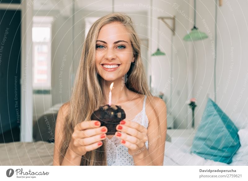 Junge schöne blonde Frau im Bett, die eine Kerze in einem Schokoladenmuffin bläst. Schlafzimmer Porträt Muffin Kuchen blasen Geburtstag Lebensmittel Fressen