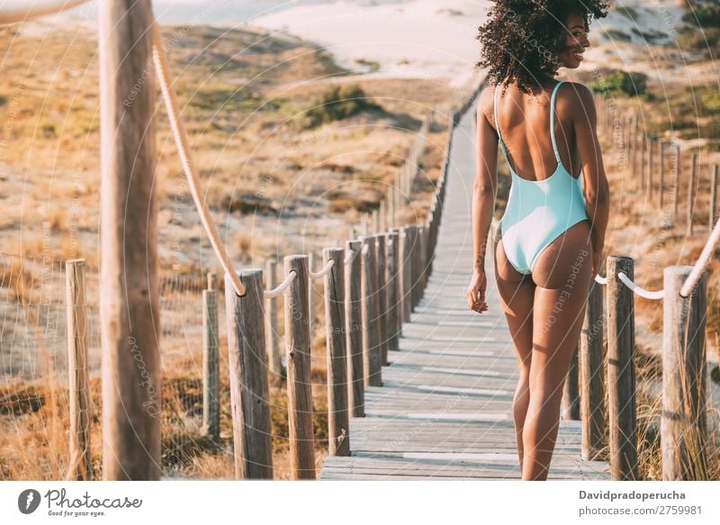 Junge Frau im Badeanzug, die in einer Holzsteg am Strand spazieren geht. Bikini Schwimmsport Anzug schwarz Brücke Hintergrundbild Küste krause Haare