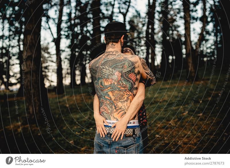 Junger Mann mit Tattoos umarmt die Dame im Wald. Paar Umarmen Schnappschuss Park ohne Hemd Typ Jugendliche Schickimicki umarmend Frau romantisch Kunst