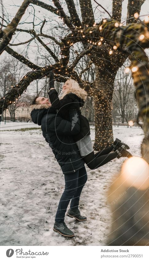 Lächelndes Paar, das Spaß auf der Straße in der Nähe eines Baumes mit Lichterketten hat. Spaß haben Weihnachten & Advent Typ Dame Skijacke Glück Hand Holz