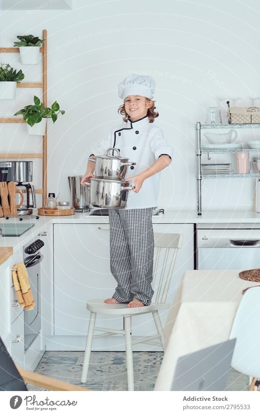 Lächelnder Junge mit Kochmütze hält Töpfe auf Stuhl in der Küche. Topf Küchenchef Kind Hut kochen & garen modern heimwärts Glück Licht vorbereitend Fröhlichkeit