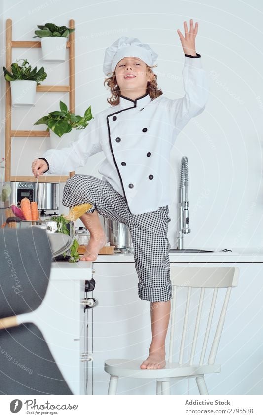 Junge mit Kochmütze mit erhobener Hand in der Nähe des Topfes auf Stuhl in der Küche. erhöht Küchenchef Kind Gemüse Hut kochen & garen modern lustig heimwärts