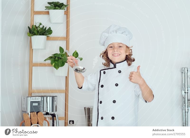 Junge mit Kochmütze hält Schöpfkelle in der Nähe des Mundes in der Küche. Schöpflöffel Küchenchef Kind Topf Verkostung Lebensmittel Hut kochen & garen Metall