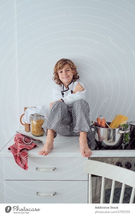 Junge sitzt in der Nähe von Töpfen auf der Elektrofritteuse in der Küche. Koch Topf Küchenchef Kind Gemüse Hut Herd & Backofen kochen & garen modern lustig