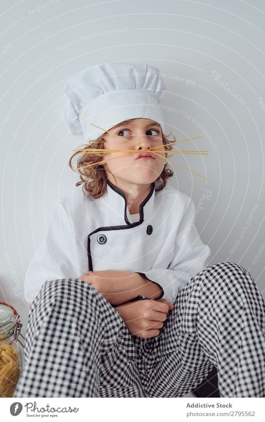 Junge mit Kochmütze, der in der Küche einen Schnurrbart aus Makkaroni macht. Oberlippenbart Küchenchef Kind Hut Spätzle regenarm machen kochen & garen modern