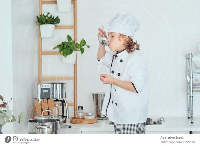 Junge mit Kochmütze hält Schöpfkelle in der Nähe des Mundes in der Küche. Schöpflöffel Küchenchef Kind Topf Verkostung Lebensmittel Hut kochen & garen Metall