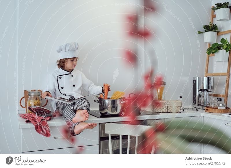 Junge mit Kochmütze und Buch in der Nähe von Töpfen auf der Elektrofritteuse in der Küche. Topf Küchenchef Kind Gemüse Hut Lautstärke lesen Herd & Backofen