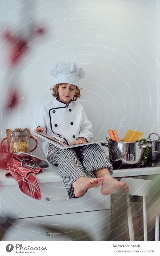 Junge mit Kochmütze und Buch in der Nähe von Töpfen auf der Elektrofritteuse in der Küche. Topf Küchenchef Kind Gemüse Hut Lautstärke lesen Herd & Backofen