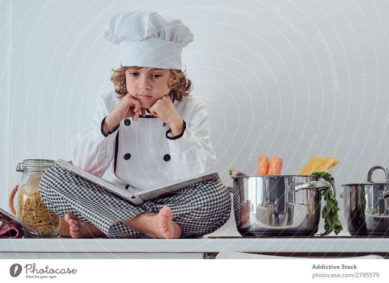 Junge mit Kochmütze und Buch in der Nähe von Töpfen auf der Elektrofritteuse in der Küche. Topf Küchenchef Kind Gemüse Hut Lautstärke lesen Herd & Backofen