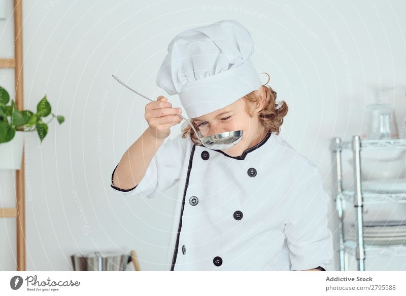 Junge mit Kochmütze hält Schöpfkelle in der Nähe des Mundes in der Küche. Schöpflöffel Küchenchef Kind Topf Verkostung Lebensmittel Hut kochen & garen Metall