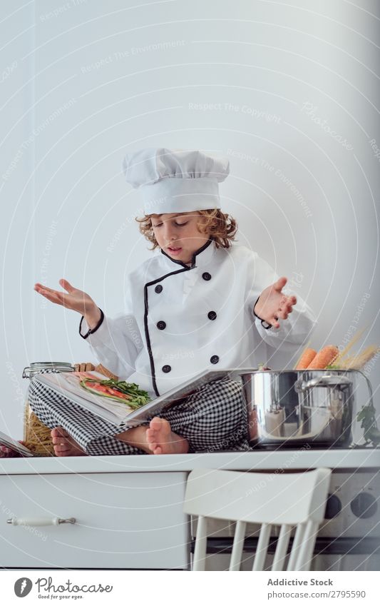 Junge mit Kochmütze und Buch in der Nähe von Töpfen auf der Elektrofritteuse in der Küche. Topf Küchenchef Kind Gemüse Hut Lautstärke lesen Herd & Backofen