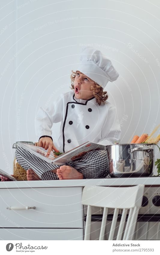 Junge mit Kochmütze und Buch in der Nähe von Töpfen auf der Elektrofritteuse in der Küche. Topf Küchenchef Kind Gemüse Hut Lautstärke lesen Herd & Backofen