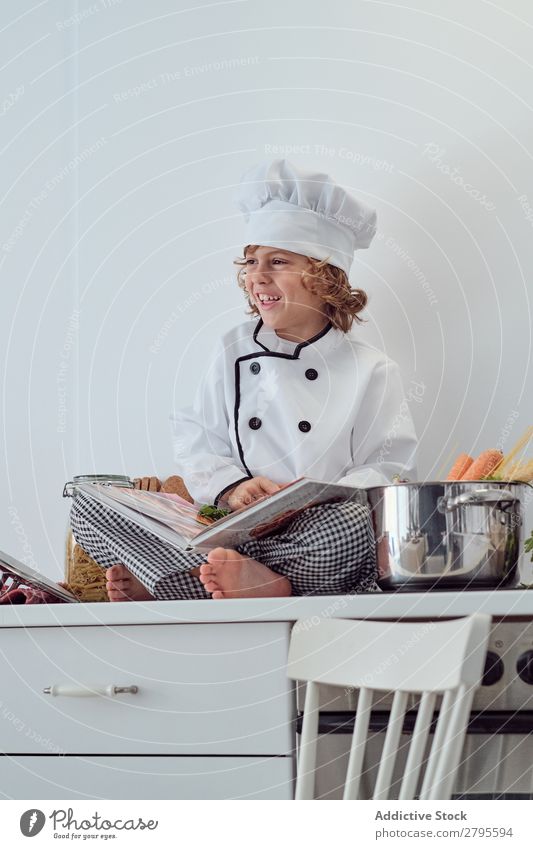 Junge mit Kochmütze und Buch in der Nähe von Töpfen auf der Elektrofritteuse in der Küche. Topf Küchenchef Kind Gemüse Hut Lautstärke lesen Herd & Backofen