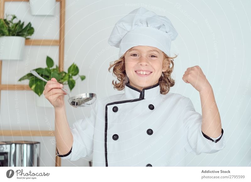 Junge mit Kochmütze hält Schöpfkelle in der Nähe des Mundes in der Küche. Schöpflöffel Küchenchef Kind Topf Verkostung Lebensmittel Hut kochen & garen Metall