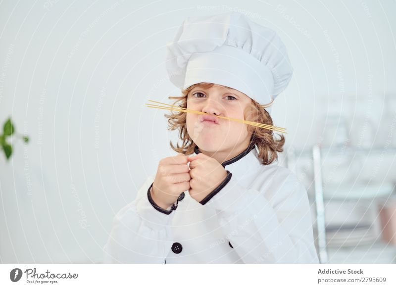 Junge mit Kochmütze, der in der Küche einen Schnurrbart aus Makkaroni macht. Oberlippenbart Küchenchef Kind Hut Spätzle regenarm machen kochen & garen modern