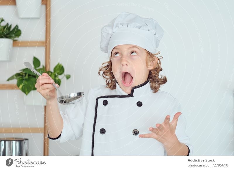 Junge mit Kochmütze hält Schöpfkelle in der Nähe des Mundes in der Küche. Schöpflöffel Küchenchef Kind Topf Verkostung Lebensmittel Hut kochen & garen Metall