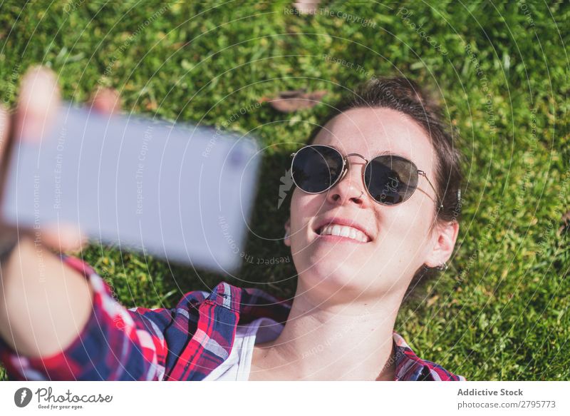 Oben die junge lächelnde Hipsterfrau, die an einem sonnigen Tag im Park auf Gras liegt und dabei mit dem Handy ein Selfie nimmt. obere Ansicht Junge Frau