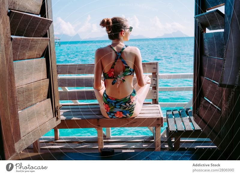 Frau im Bikini sitzt auf einer Bank in der Nähe des Wassers. Meer Jamaika Sonnenbrille Badeanzug Sitz blau tropisch exotisch sitzen Accessoire Sommer dünn Dame