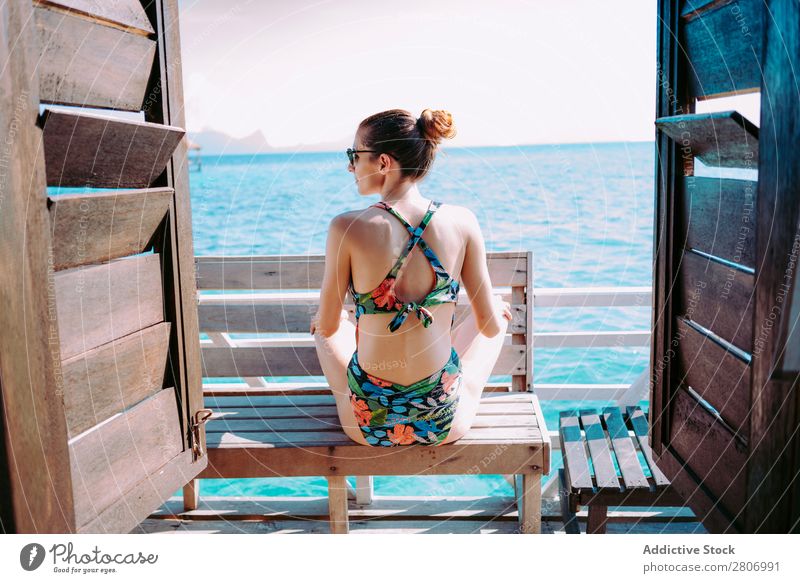 Frau im Bikini sitzt auf einer Bank in der Nähe des Wassers. Meer Jamaika Sonnenbrille Badeanzug Sitz blau tropisch exotisch sitzen Accessoire Sommer dünn Dame