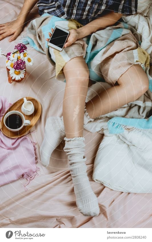 Weiße Frau in weißen Socken sitzt im Bett und hält ein Smartphone. PDA Lifestyle heimwärts Erwachsene Beautyfotografie Bettwäsche Getränk Decke Bettdecke