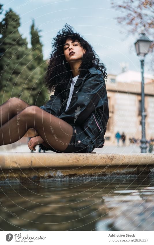 Junge Frau, die am Springbrunnen posiert. attraktiv Großstadt modisch lockig brünett Jacke Wasser Blick in die Kamera Mode Jugendliche schön hübsch Straße Model