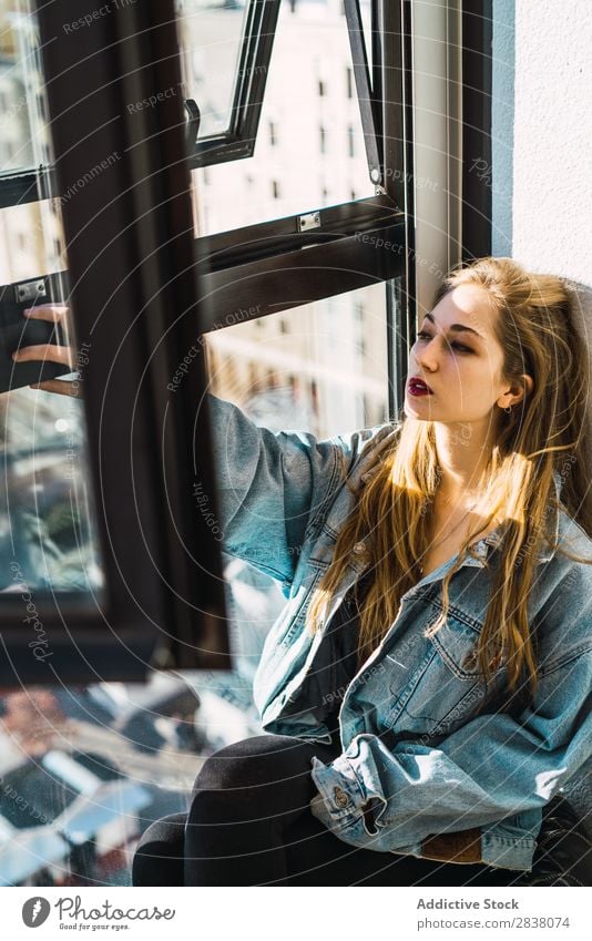 Stylisches Mädchen sitzt am Fenster im Licht. Frau Stil sitzen Körperhaltung Bekleidung Lippen genießen lange Haare Lippenstift träumen Jeansstoff Sonnenlicht