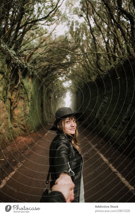 Junge stilvolle Frau hält Hand auf dem Weg zwischen hohen Mauern und Bäumen. Händchenhalten mir folgen Baum Wand Gasse Fußweg dunkel Stil elegant Lederjacke Hut