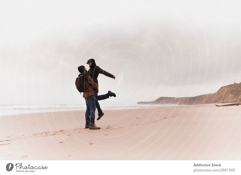 Ein Paar küsst sich auf dem Sandufer in der Nähe des Wassers. Strand Küssen kalt Küste Wärme anhaben Rucksack Hand Halt Mann Frau Hut Meer Hügel Jugendliche