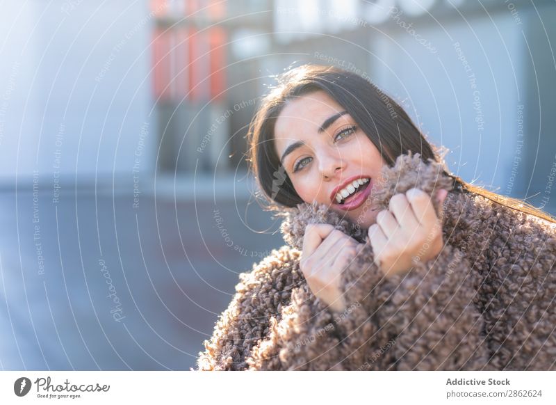 Fröhliche Frau, die auf der Straße in einen Mantel gehüllt ist. heiter Verpackung Großstadt Stil Jugendliche Sonnenstrahlen Tag genießen trendy Mode Outfit