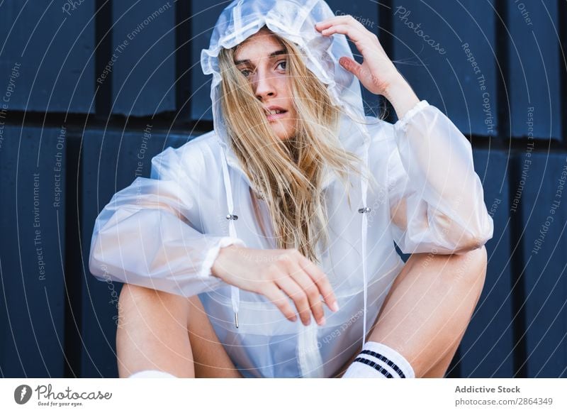 Charmante, stylische Frau im Regenmantel, die an der Wand sitzt. blond charmant Straße Dame genießen Jeans Shorts Kniestrumpf Stil sitzen Jugendliche Model