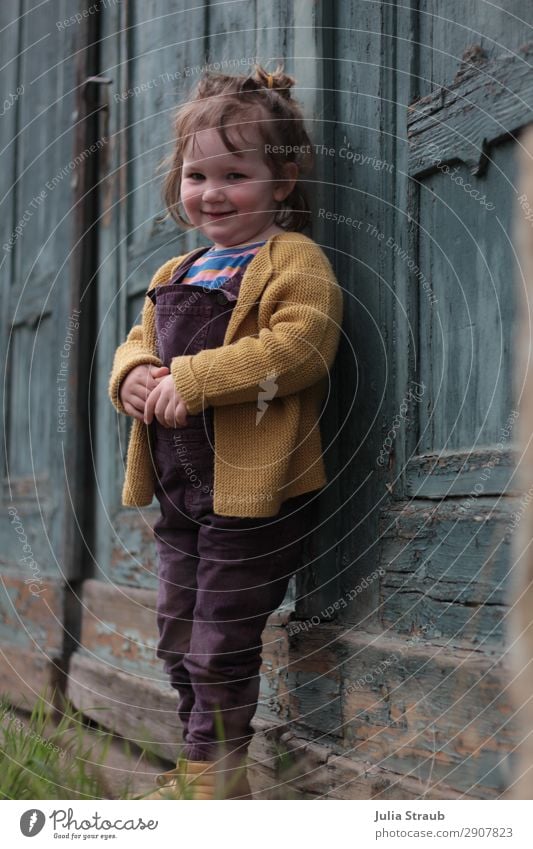 Mädchen alte Tür Holz feminin Kleinkind 1 Mensch 1-3 Jahre Gebäude Latzhose Strickjacke brünett kurzhaarig Pony Zopf Lächeln Scham verlegen Holztor abblättern