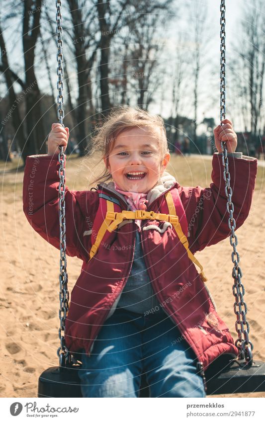 Kleines lächelndes glückliches Mädchen schaukelt in einem Park an einem sonnigen Frühlingstag. Kind schaut in die Kamera und trägt rote Jacke Lifestyle Freude