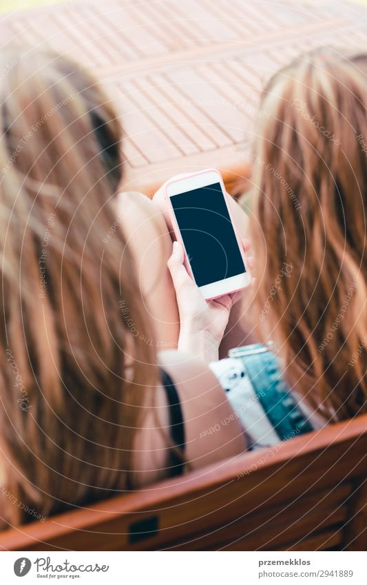 Junge Frauen, die Mobiltelefone benutzen und Musikclips ansehen, SMS schreiben, Nachrichten versenden. Teenager, die Smartphones benutzen, sitzen auf einer Bank im Freien. Verwendung von Technologiegeräten. Mädchen tragen Sommerkleidung