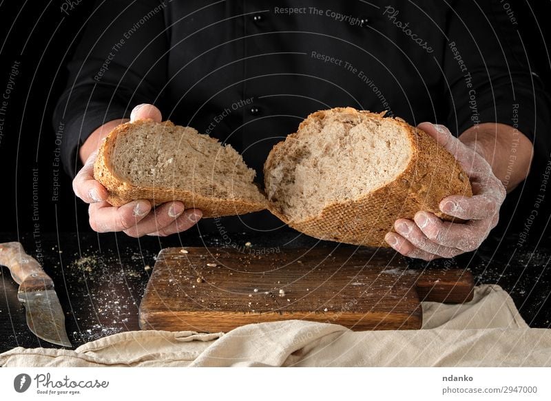 Chefkoch hält gebrochenes halbrundes Roggenbrot in der Hand. Brot Ernährung Messer Tisch Küche Mensch Finger Holz machen dunkel frisch heiß braun schwarz weiß