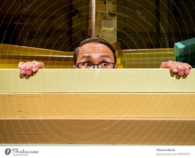 Junger Mann schaut neugierig über eine Absperrung Innenaufnahme 1 Mensch Kunstlicht Porträt Blick nach vorn Blick in die Kamera Brille Zaun Barriere Holzzaun