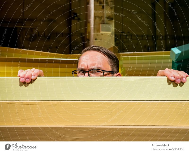 Junger Mann schaut neugierig über eine Absperrung Innenaufnahme 1 Mensch Kunstlicht Porträt Blick nach vorn Blick in die Kamera Brille Zaun Barriere Holzzaun