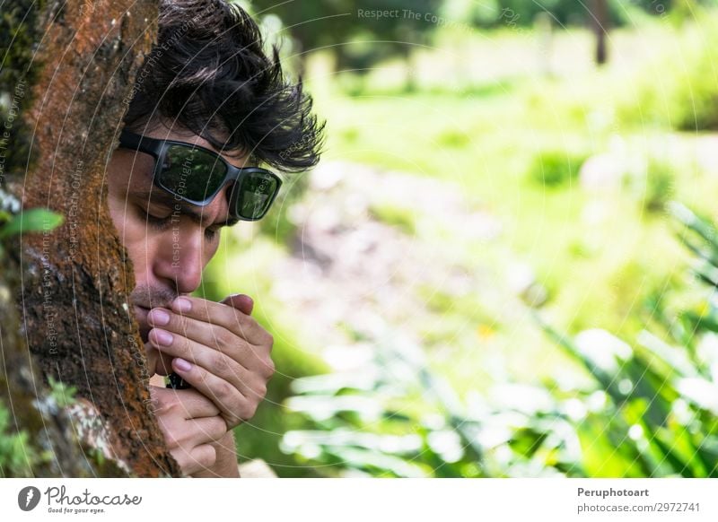 Junger Mann, der eine Zigarre hinter einem Baum im Wald anzündet. Alkohol Lifestyle Reichtum Gesicht Erholung Mensch maskulin Erwachsene Park Sonnenbrille alt