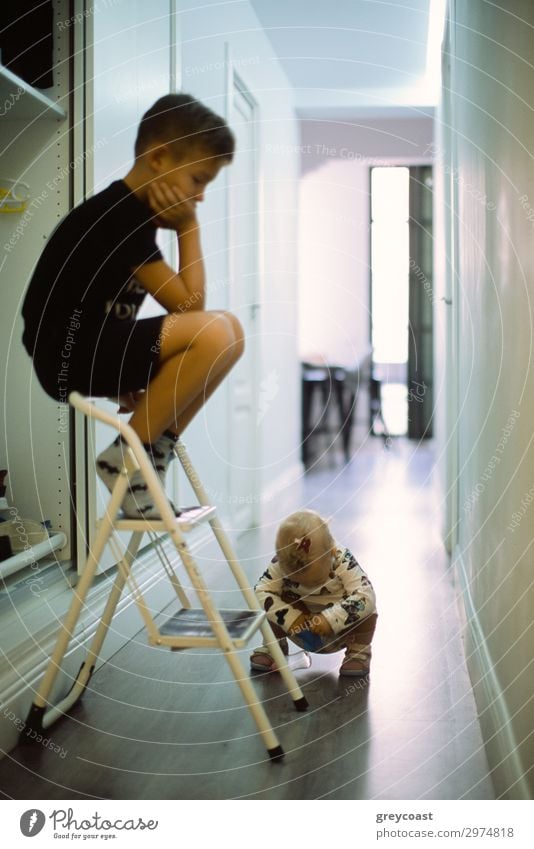 Ein kleines Mädchen studiert etwas auf den Hüften, während ihr älterer Bruder sie von oben auf der Treppe sitzend beobachtet Leben Freizeit & Hobby Spielen Kind