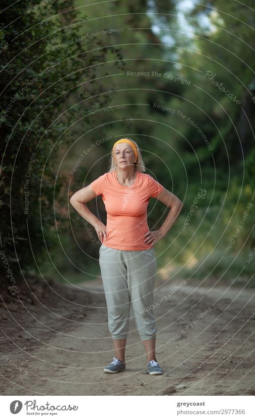 Eine Frau mittleren Alters steht auf einem Parkweg, die Hände in die Hüften gestemmt, müde von einer Joggingrunde Sport Mensch feminin Erwachsene