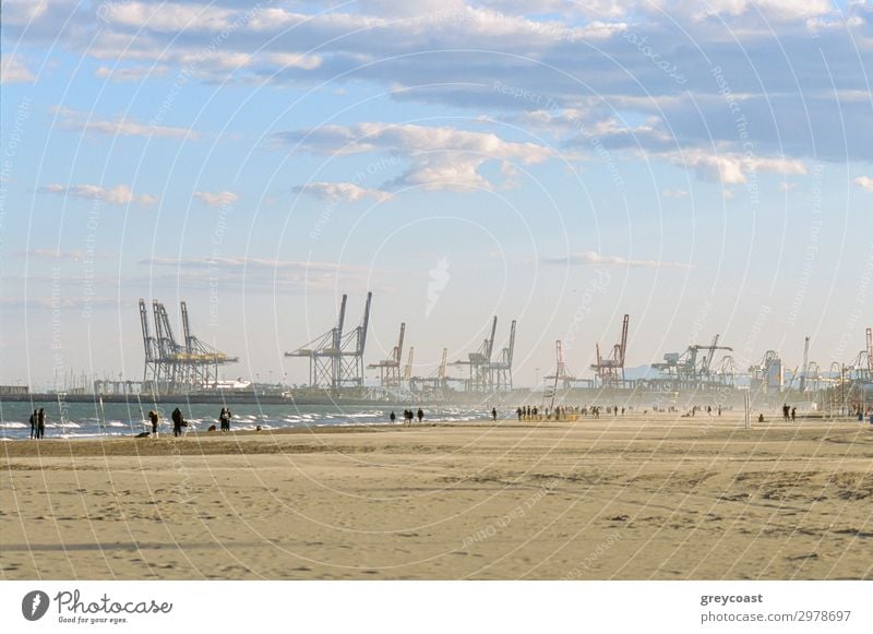 Menschen spazieren am Strand von Valencia im Winter, Spanien. Szene mit Industriehafen und Containerkränen Meer Himmel Wellen Küste Binnenschifffahrt Hafen