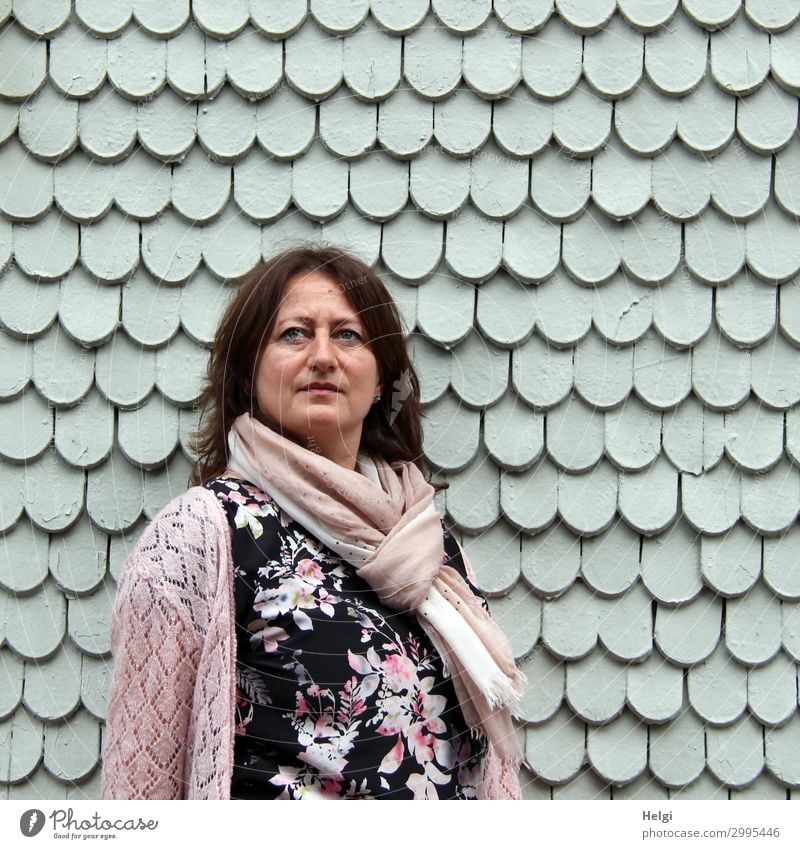 Frau mit dunklen langen Haaren , buntem Kleid, rosa Strickjacke und rosa Schal steht vor einer grauen Wand aus Holzschindeln Mensch feminin Erwachsene 1