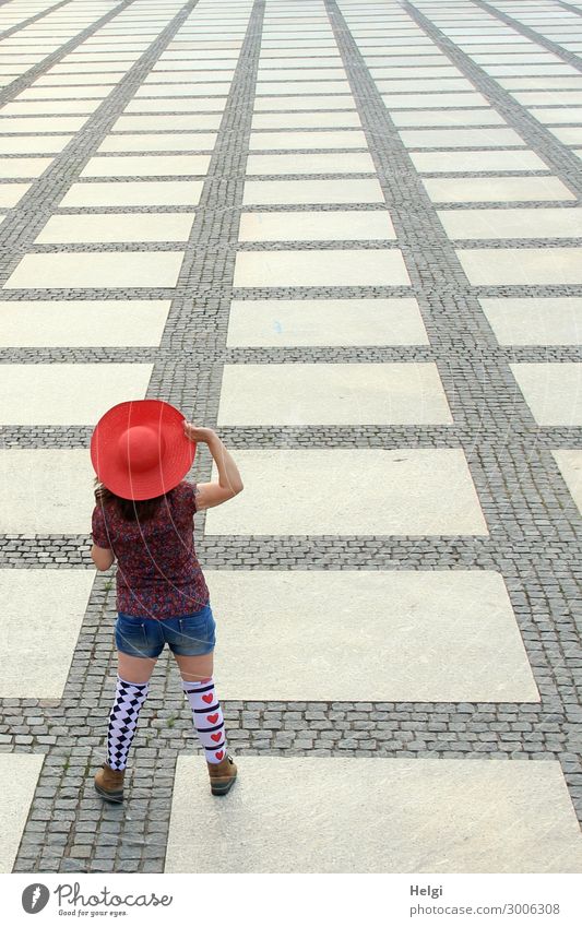 Rückansicht einer Frau mit rotem Hut, kurzer Jeans, Bluse und gemusterten Strümpfen steht auf einem großen Platz mit gemustertem Belag Mensch feminin Erwachsene