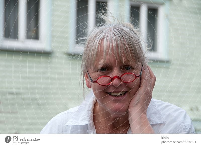 Reife Frau mit roter Brille auf der Nase, Ponyfrisur, grauen Haaren, steht vor weißem Haus mit Fenstern. Weiblicher Senior, mit Schmerzen an Ohr oder Zahn hält die Hand schützend an die Backe vom Gesicht. Dame mit erfreutem, überraschtem Gesichtsausdruck.