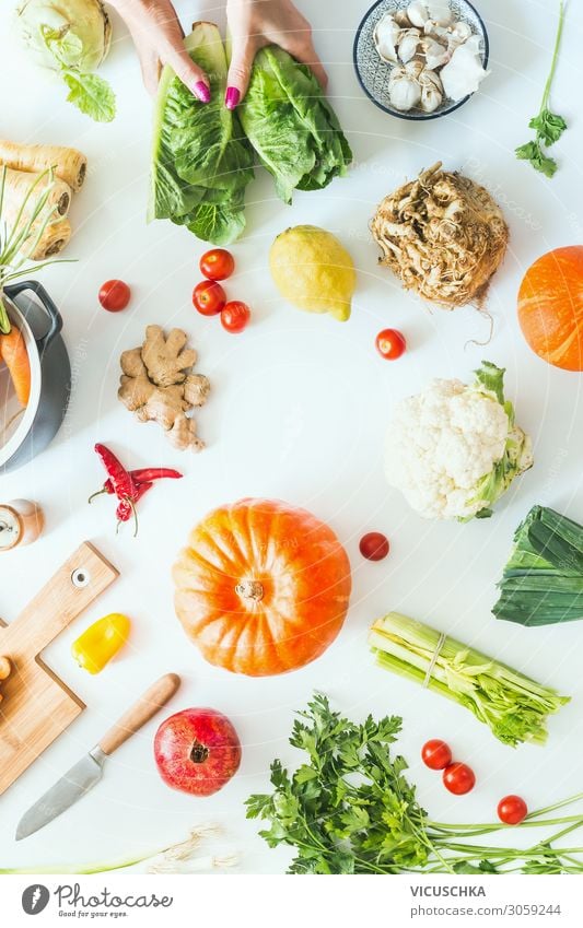 Weibliche Hände halten Salat auf weißem Tischhintergrund mit Kürbissen und verschiedenen Gemüsesorten der Saison, Topf, Schneidebrett und Messer. Kochen der Ernte. Flach liegend. Gesunde Lebensweise und Ernährung. Kohlenhydratarm