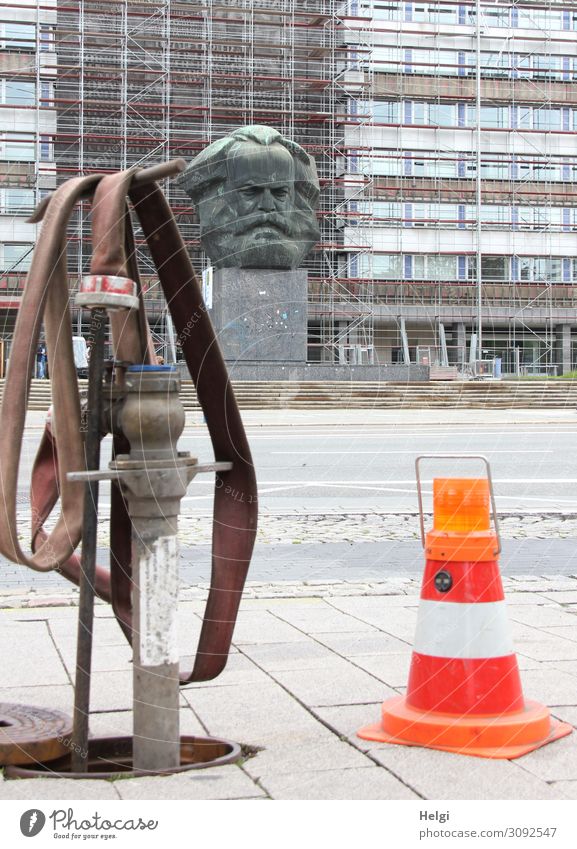 Hydrant mit Wasserschlauch und Absperrkegel mit Warnleuchte steht am Straßenrand, im Hintergrund das Karl-Marx-Denkmal vor einer Häuserfassade in Chemnitz Stadt