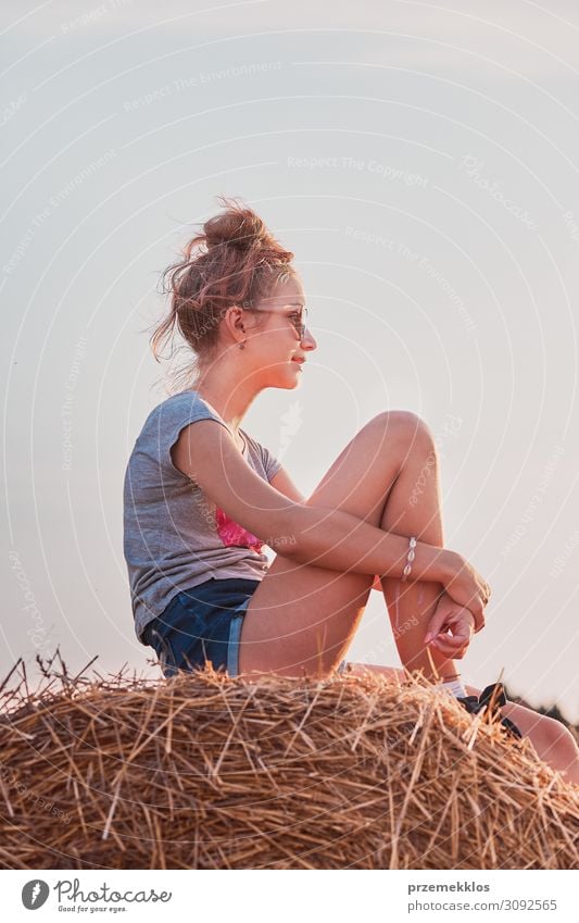 Teenager-Mädchen sitzt bei Sonnenuntergang auf einem Heuballen und entspannt sich, während sie den Sommer auf dem Dorf verbringt. Unverfälschte Menschen, echte Momente, authentische Situationen