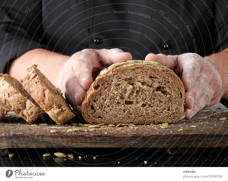 Koch in schwarzer Uniform schneidet ein Stück Brot ab. Ernährung Essen Mittagessen Abendessen Tisch Küche Mann Erwachsene Hand Holz dunkel frisch lecker