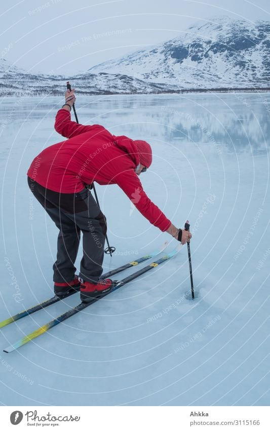 Mann mit roter Jacke, roten Schuhen und roter Mütze auf Skiern prüft mit einem Skistock die Eisdicke eines Sees Wassersport Wintersport Wissenschaften Studium