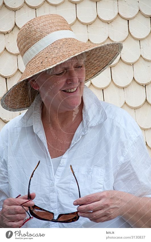 Reife Frau mit Strohhut auf dem Kopf, heller, weißer Bluse und Sonnenbrille haltend in der Hand, steht im Urlauf vor einem alten Haus mit weissen Schindeln. Dame steht beim Ausflug in der Altstadt vor heller Wand und lächelt glücklich seitwärts nach unten.