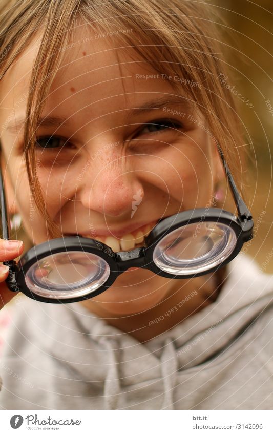 Lachendes, fröhliches Mädchen zieht dicke Hornbrille; Scherzartikel mit hoher Dioptrin aus Spaß, Verkleidung und Freude, an Karneval; Fasching an und lacht dabei fröhlich in die Kamera.